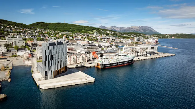 Quality Hotel Harstad, a modern building on a pier in a vibrant city harbor with a large ship docked, surrounded by hills and mountains.