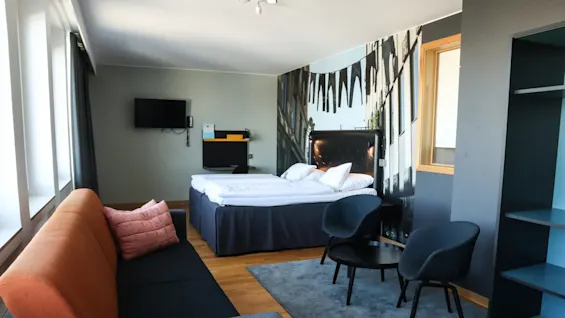 En säng är prydligt bäddad i ett hotellrum, med en väggmålning av hängande jeans i bakgrunden. En soffa och stolar kompletterar den moderna inredningen.