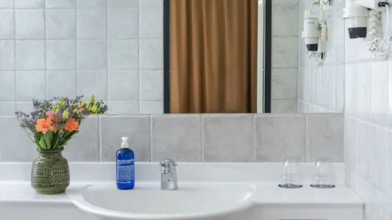 A bathroom sink holds a green vase with colorful flowers and a blue soap bottle reading "MALIN+GOETZ." Nearby, two upside-down glasses rest on the white counter near a reflective mirror.