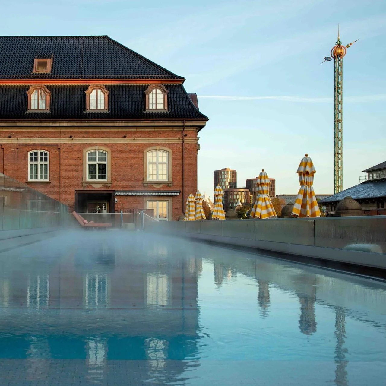 A misty pool reflects a historic brick building with dormer windows, surrounded by closed yellow-striped umbrellas in an urban setting with a tall, decorative tower in the background.