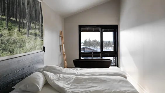 A bed with white linens rests in a minimalistic room. A black bathtub is by the window with snowy trees visible outside. A wall features a large forest-themed print.