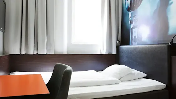 Bed positioned beside a window with closed curtains, adjacent to a wall displaying a movie projector image; a desk with a chair is in the foreground.