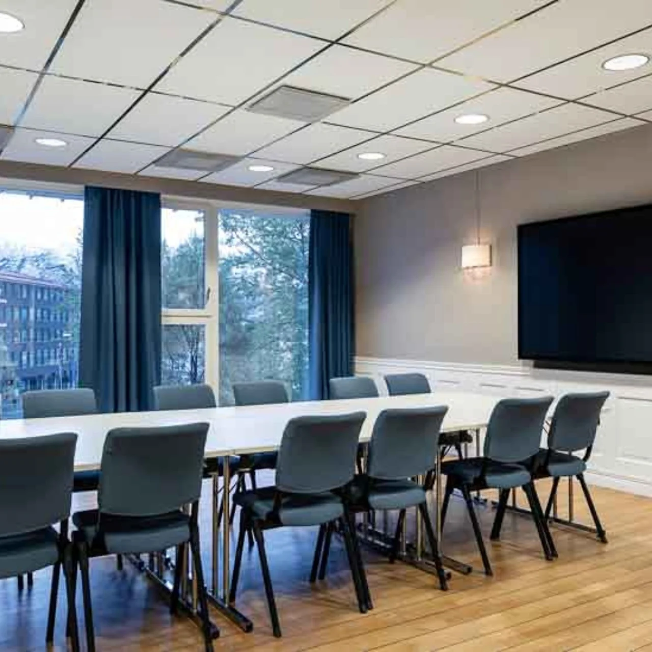 A conference table is surrounded by chairs, facing a large black screen on the wall. Large windows with blue curtains overlook trees and buildings outside.