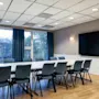 Et konferencenbord omgives af stole og vender mod en stor sort skærm på væggen. Store vinduer med blå gardiner giver udsigt til træer og bygninger udenfor.