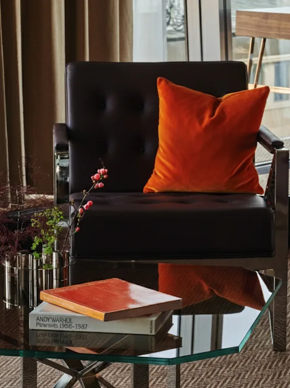 En stilfull lounge med en svart läderfåtölj, en orange kudde och ett soffbord i glas med böcker, inklusive en med titeln ANDY WARHOL 1958-1987.