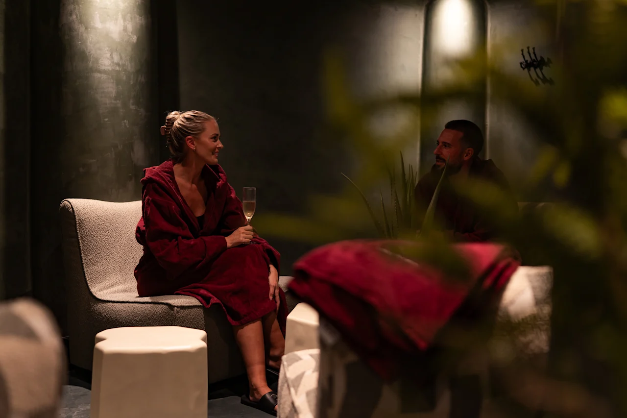 Kaksi kylpytakkiin pukeutunutta henkilöä rentoutuu ja keskustelee hämärässä oleskelutilassa, toisella on samppanjalasi.