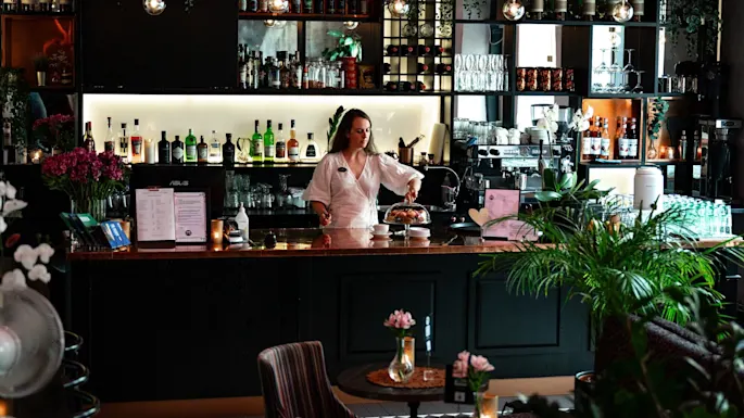 A bartender arranges pastries under a glass dome in a cozy bar. Bottles and glasses neatly line the shelves behind her, surrounded by lush plants and soft lighting.