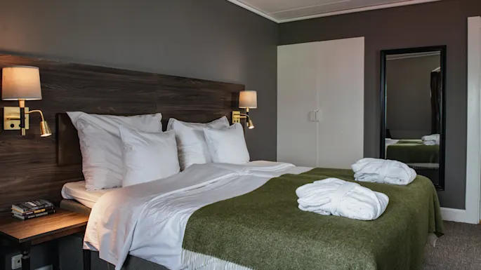 En pent oppredd seng med hvite puter og et grønt teppe står mot et mørkt trehodegjerde. Vegglamper lyser opp rommet, med et speil og nattbord i nærheten.