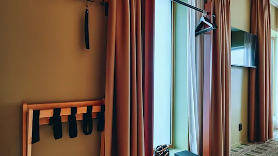 Et hotellrom med klesstativ, bagasjestativ og gardiner, designet for gjestenes komfort og organisering.