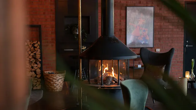 Peis stråler varme, omgitt av stoler i et koselig rom med synlige teglvegger. En kurv med ved og et innrammet bilde gir rustikk atmosfære.