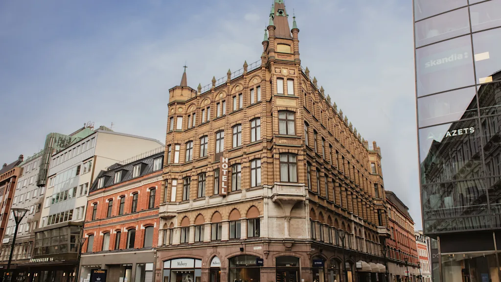Home Hotel Baltzar. A grand, ornate brick building with a spire stands on a city street, with other buildings nearby. Text: HOTELL.