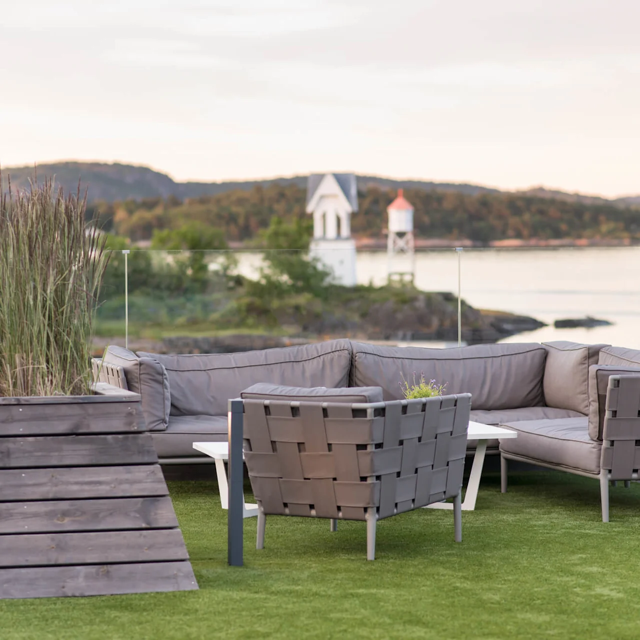 Gray outdoor furniture arranged on artificial grass, surrounded by a glass barrier. A body of water and small white structures are visible in the background.