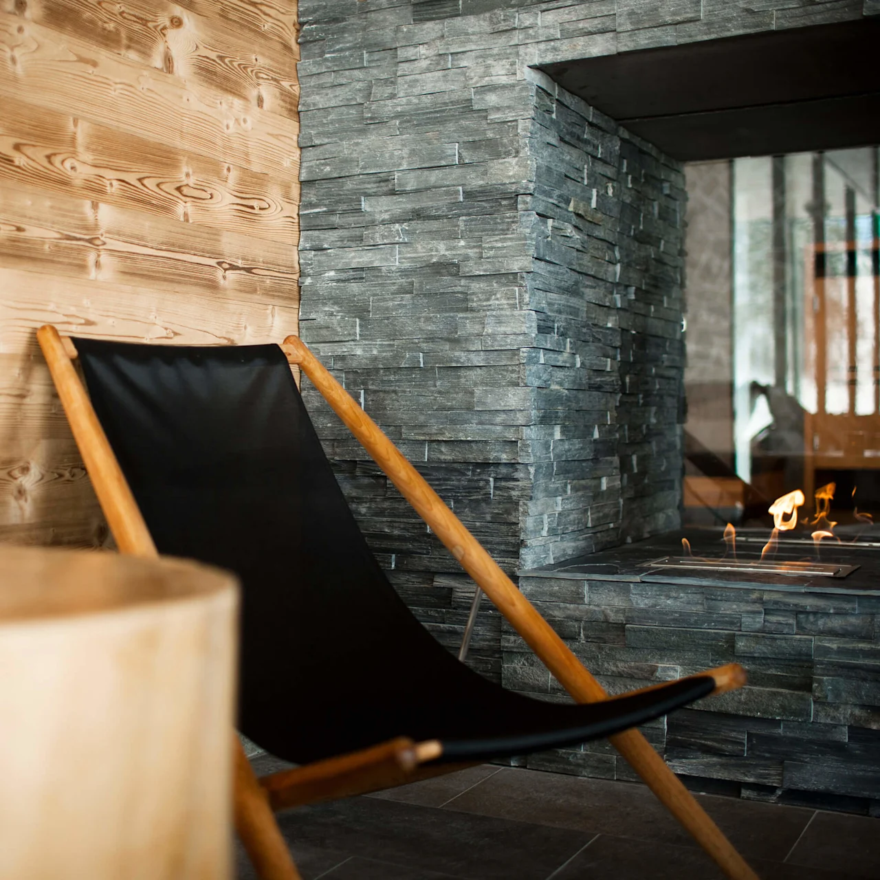 A black canvas chair near a lit fireplace, wooden and stone walls provide a cozy atmosphere.