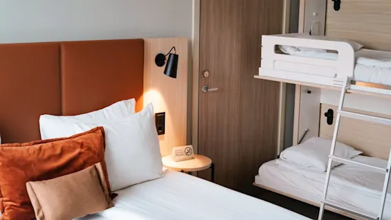 A modern room features a bed with white and rust-colored pillows, a wall light, and a bunk bed with a ladder. A "no smoking" sign is on a small table.