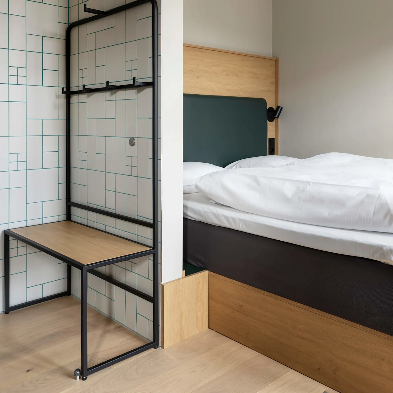 A bed with white linens sits against a beige wall. Adjacent, a black metal and wood bench is set against a patterned tile wall with geometric lines.