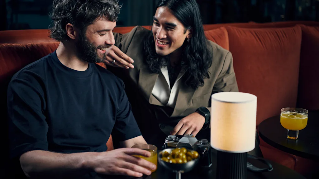 Kaksi ystävää nauttii juomia ja keskustelua viihtyisässä loungeympäristössä, jakaen naurun yhteisestä ajastaan.