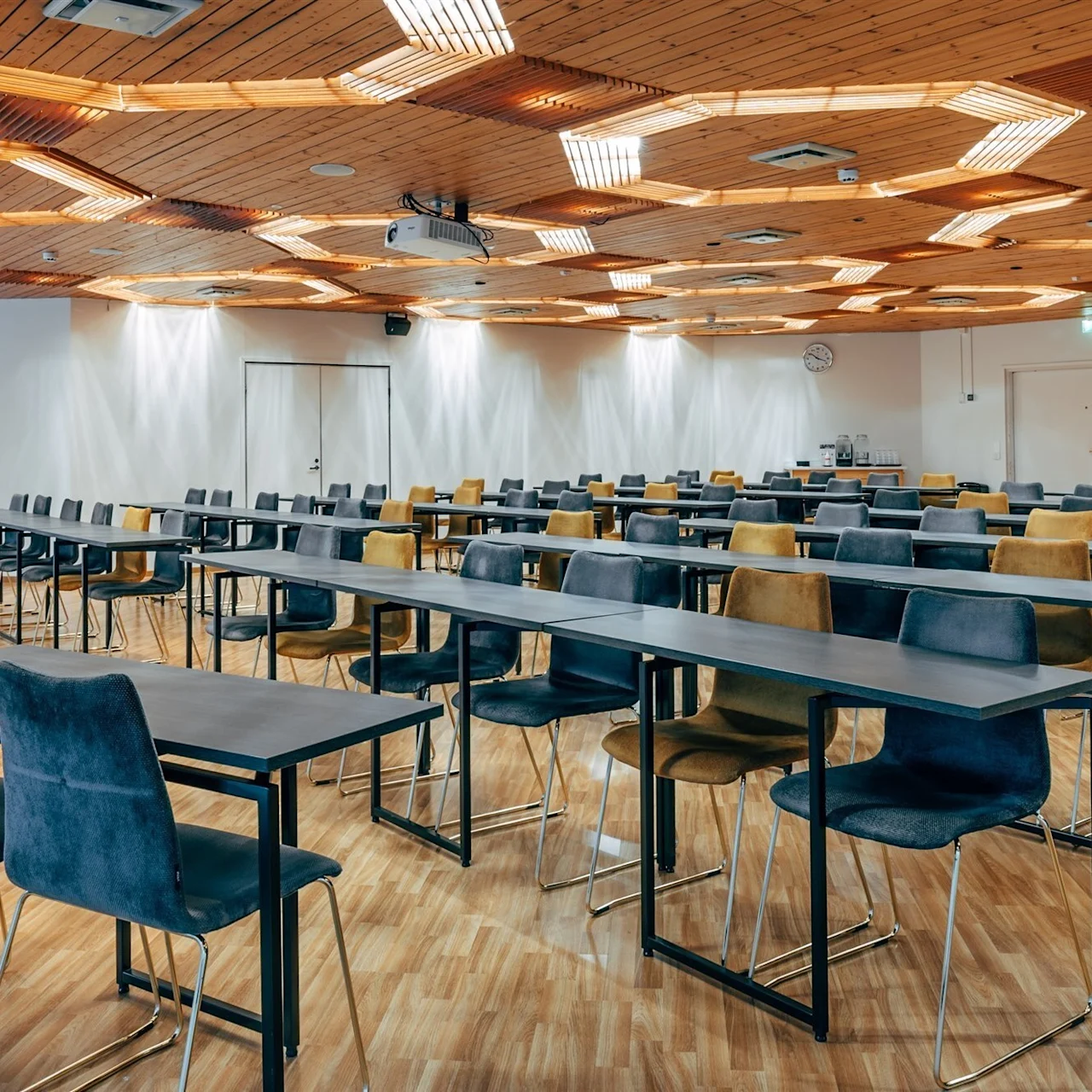 A conference room with rows of tables and chairs is set up for an event. The wooden ceiling features hexagonal lights, while the walls are plain white. Wooden flooring enhances the space.