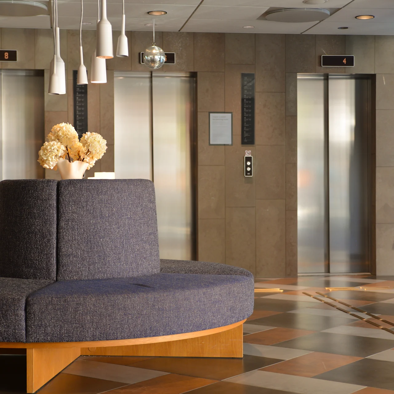 En rund, polstret bænk med en blomsterkrukke står i en lobby. Fire pendellamper hænger over. I baggrunden ses tre lukkede elevatordøre på et flisebelagt gulv.