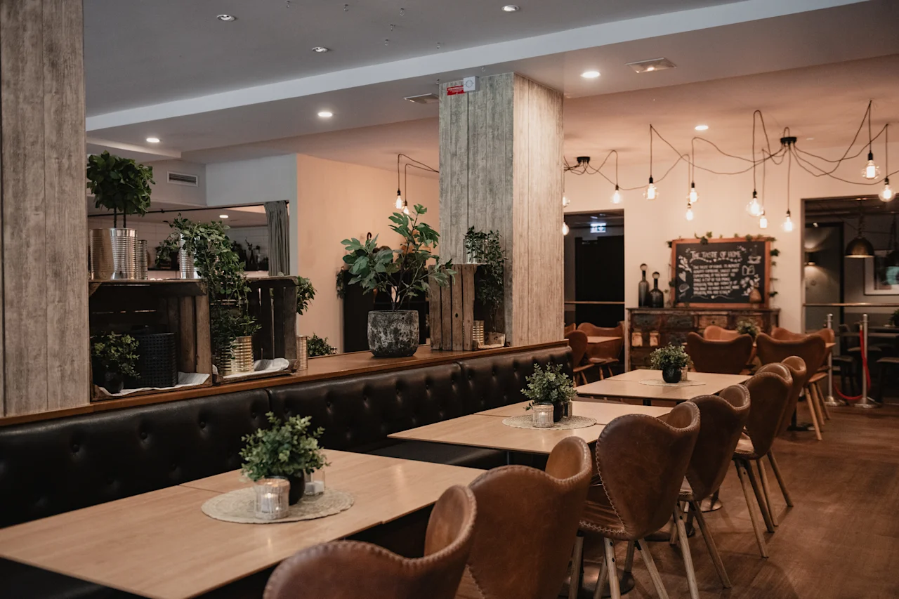 A cozy restaurant interior with wooden tables, leather chairs, and green plants. Hanging lights illuminate the space. A chalkboard reads THE TASTE OF HOME.