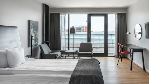 A neatly made bed sits in a modern hotel room with light wood flooring. Two armchairs and a table face large windows and a balcony, next to a sleek desk.