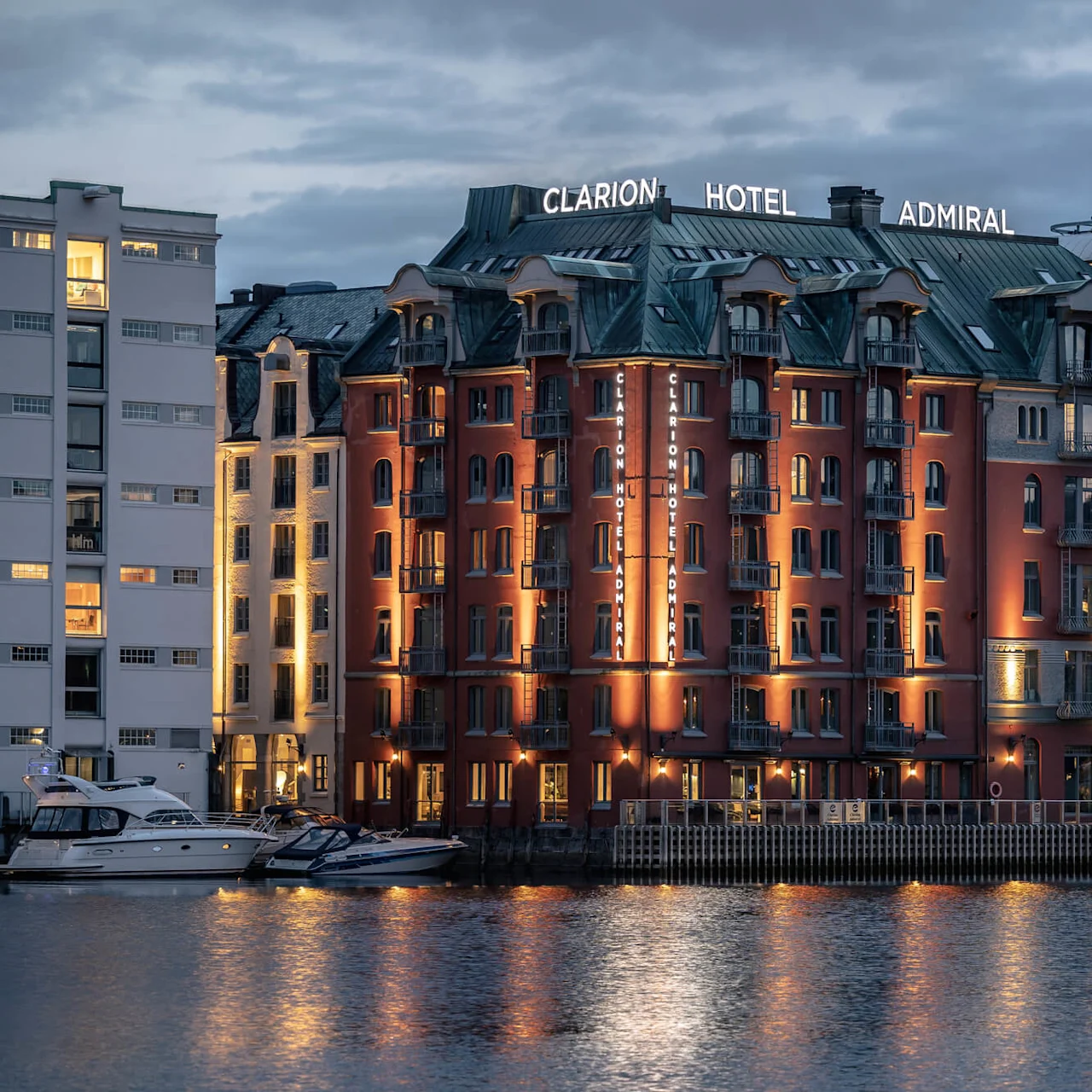 Clarion Hotel Admiral-bygget er opplyst i skumringen, med skiltet "CLARION HOTEL ADMIRAL" synlig. Det ligger ved vannkanten, omringet av moderne bygninger og båter ved kai.