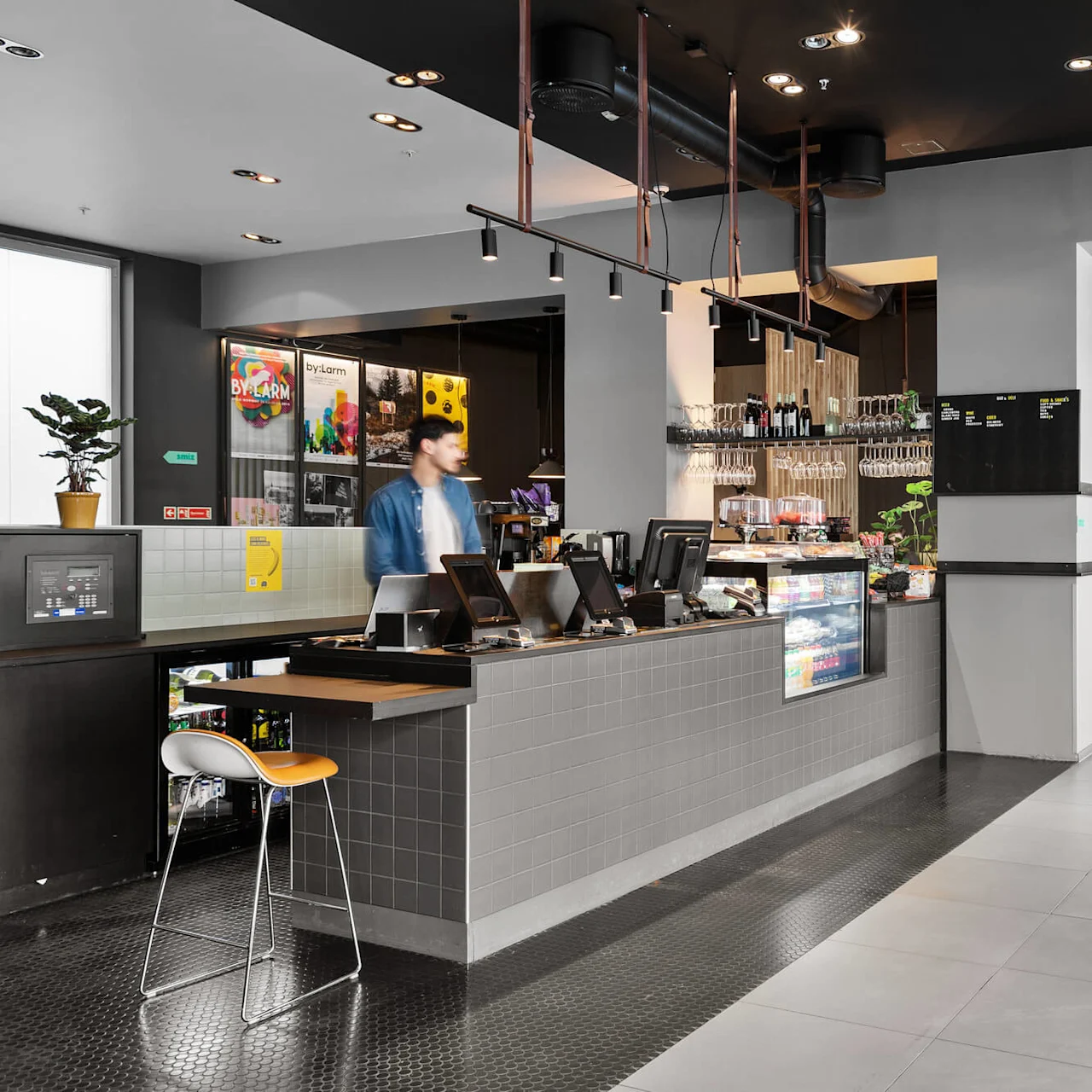 A cafe counter with a person standing, surrounded by modern decor and seating. Neon sign reads "WORKING CLASS HEROES." Floor mat text: "Good to see you" and "Pssss."