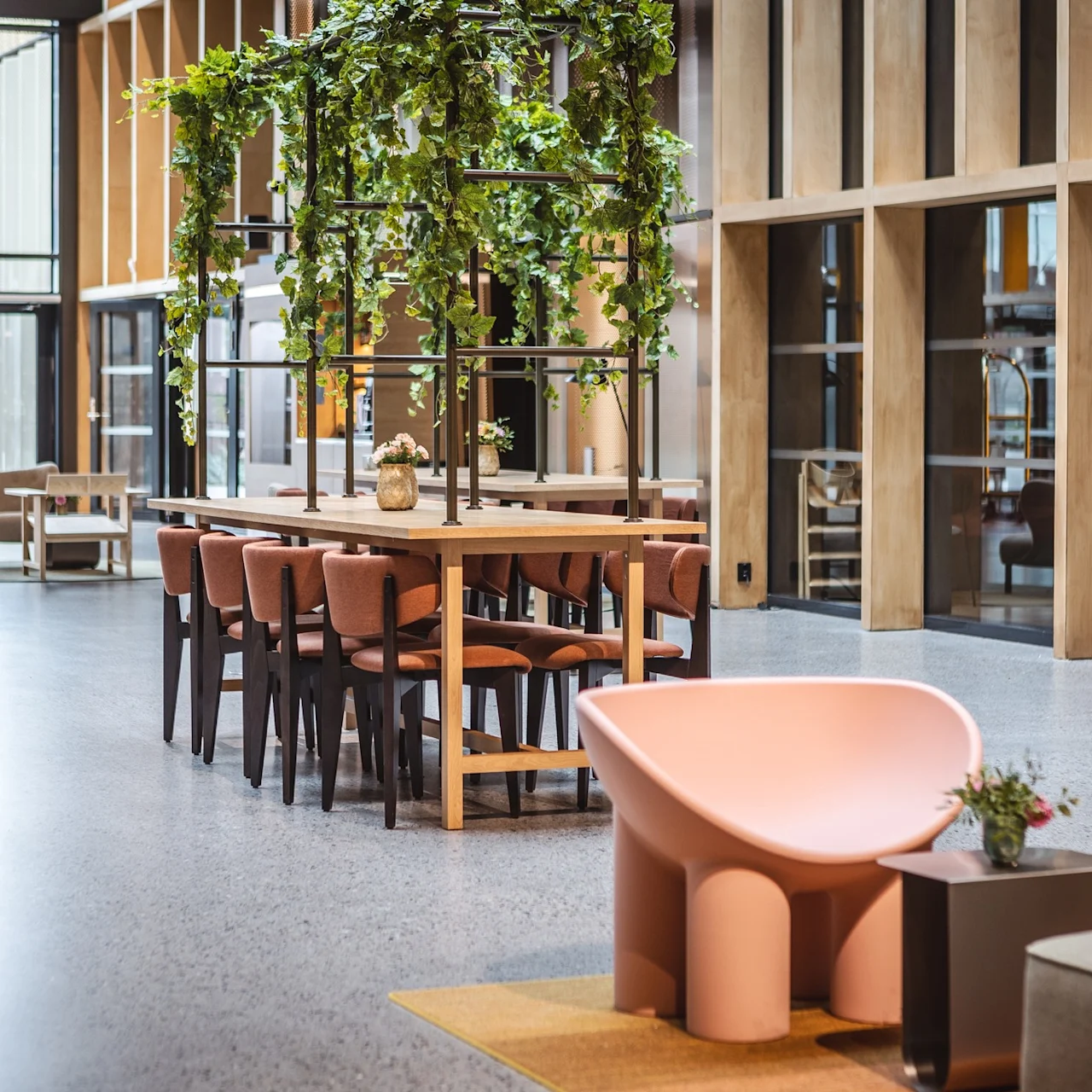 Ett långt träbord med rosa stolar är dekorerat med hängande gröna växter, beläget i en rymlig, modern lobby med stora fönster och fler sittplatser.