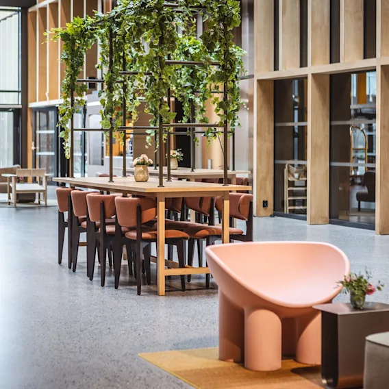 Et langt trebord med rosa stoler er dekorert med hengende grønne planter, plassert i en romslig, moderne lobby med store vinduer og flere sitteområder.