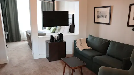 Et hotellrom med en grønn sofa, veggmontert TV og en seng i det tilstøtende rommet gjennom et vindu i veggen.