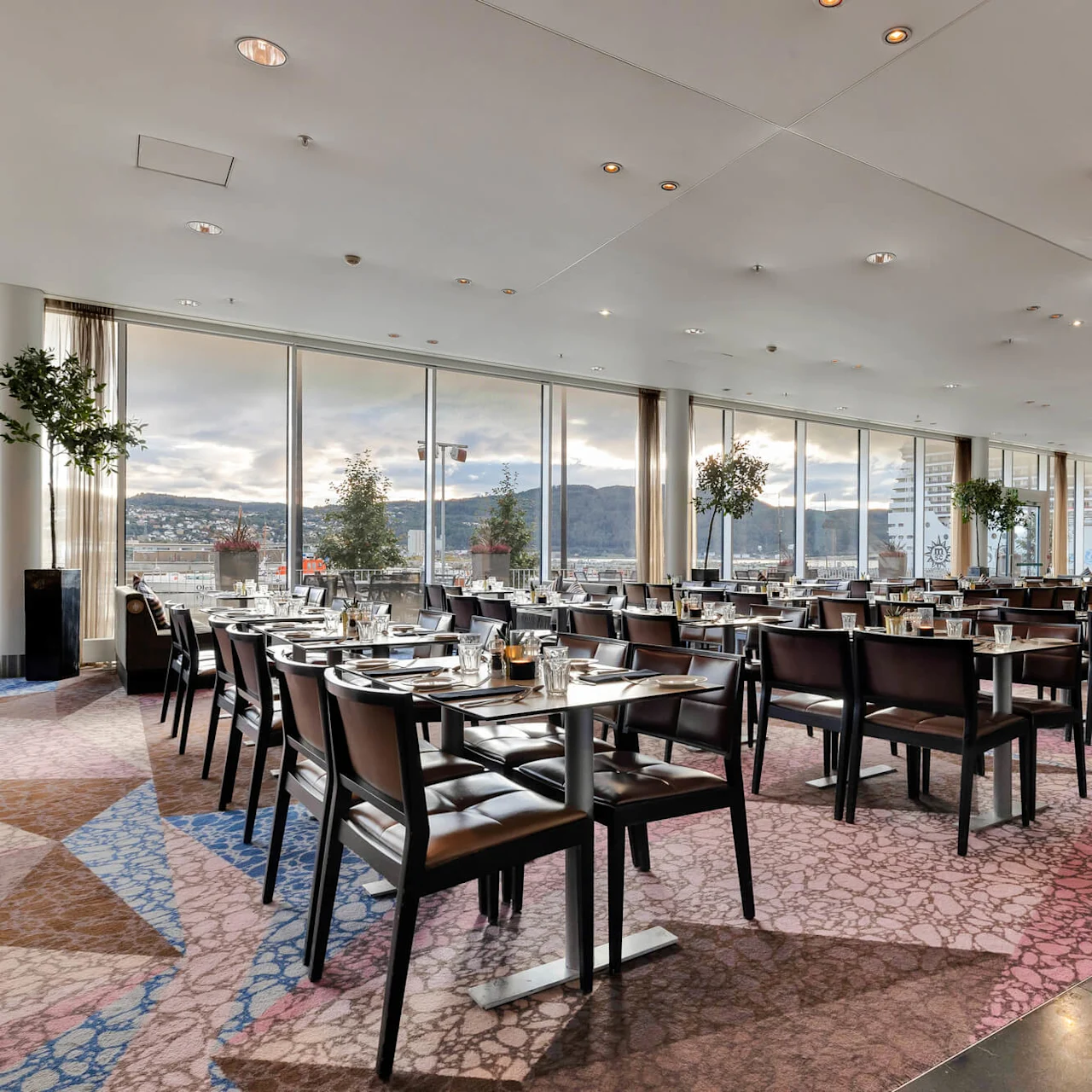 Restaurant tables set for dining, overlooking large windows with a scenic view of mountains and city buildings. The modern interior features patterned carpeting and decorative plants.