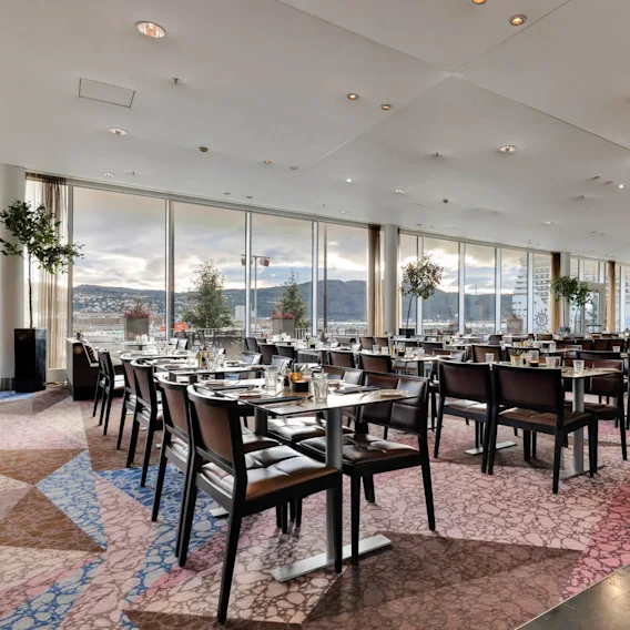 Restauranterbord dekket for middag, med utsikt over store vinduer med fjell og bybygninger. Det moderne interiøret har mønstret teppe og dekorative planter.