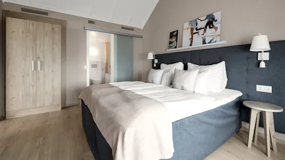En pent queen-size-seng med hvitt sengetøy og puter står mot en beige vegg, flankert av moderne hvite lamper og et trebord. Et skap og et bad er synlige.