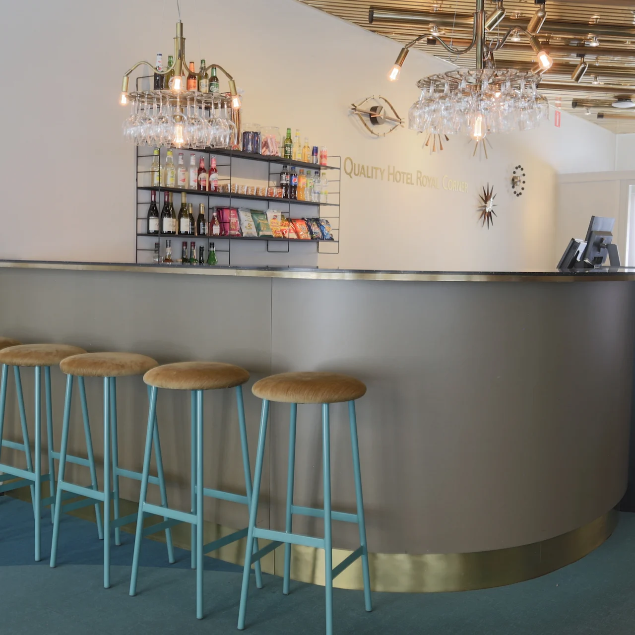 Bar counter with five stools, lit overhead shelves displaying bottles and snacks. Crystal chandeliers hang above. White wall text reads: "Quality Hotel Sarpsborg". Sign reads: "SOCIAL Welcome to The Social Bar & Bistro".