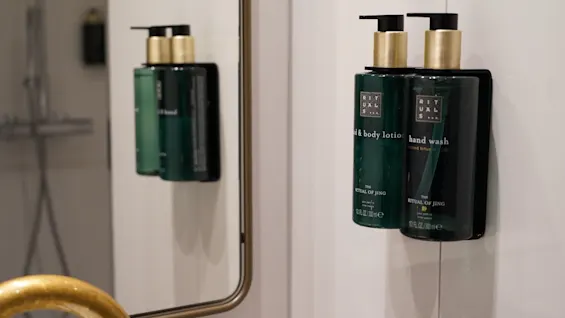Hand wash and body lotion bottles, labeled "RITUALS THE RITUAL OF JING," mounted on a bathroom wall, reflected in a mirror, next to a golden faucet.