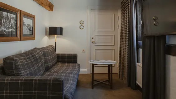 Et hyggeligt værelse med en ternet sofa, en gulvlampe og indrammede billeder. Synlige træbjælker og et lille bord fuldender det indbydende rum.