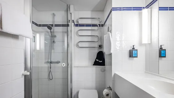A bright bathroom with white tiles and blue accents. The bathroom features a glass-enclosed shower, a wall-mounted toilet, and a modern sink. 