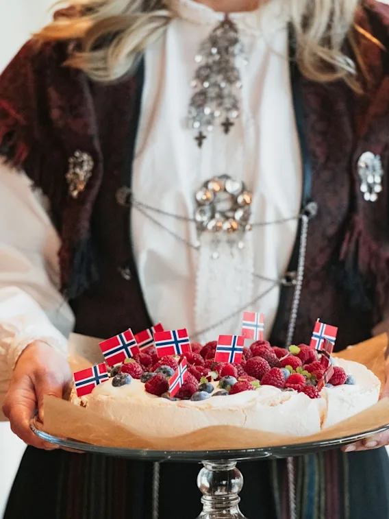 Nærbilde av en person i bunad som holder en pavlova toppet med bringebær, blåbær og små norske flagg.
