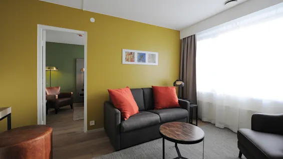 Gray sofa with red cushions sits in a living room. Nearby, a round coffee table stands on a gray rug. Large windows with curtains let in natural light.