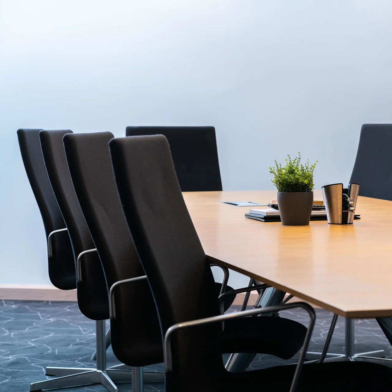 Ett träbord i konferensrummet har en krukväxt och metallkoppar, omgiven av svarta stolar i ett modernt mötesrum med matta.