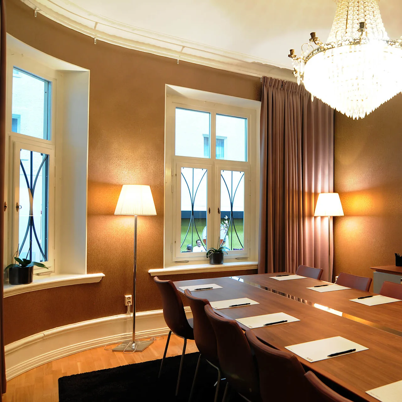 A conference room with a long table, chairs, papers and pens, bright from lamps and natural light from windows looking onto a yard.