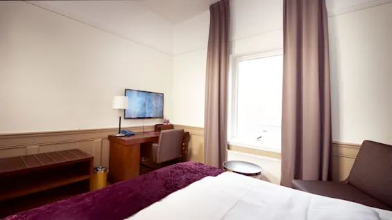 En pent oppredd seng med en burgunder kast ved fotenden, et TV på et stativ, og et vindu med trukne gardiner i et ryddig hotellrom.