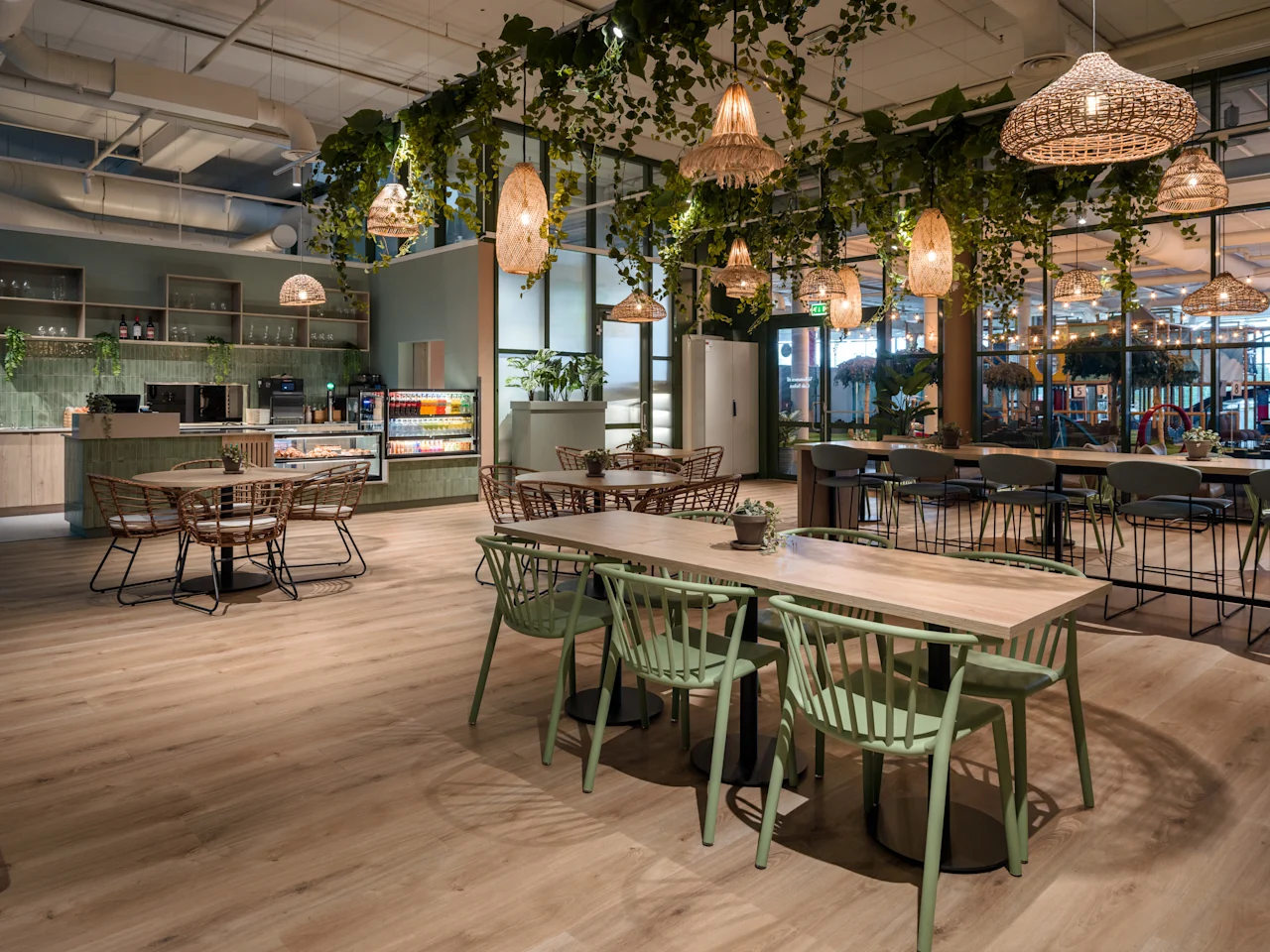 Borde og stoler er arrangert i en moderne kafé, med hengende lys og grønt over, i et romslig, godt opplyst interiør med tregulv og hyller fylt med kjøkkenartikler.
