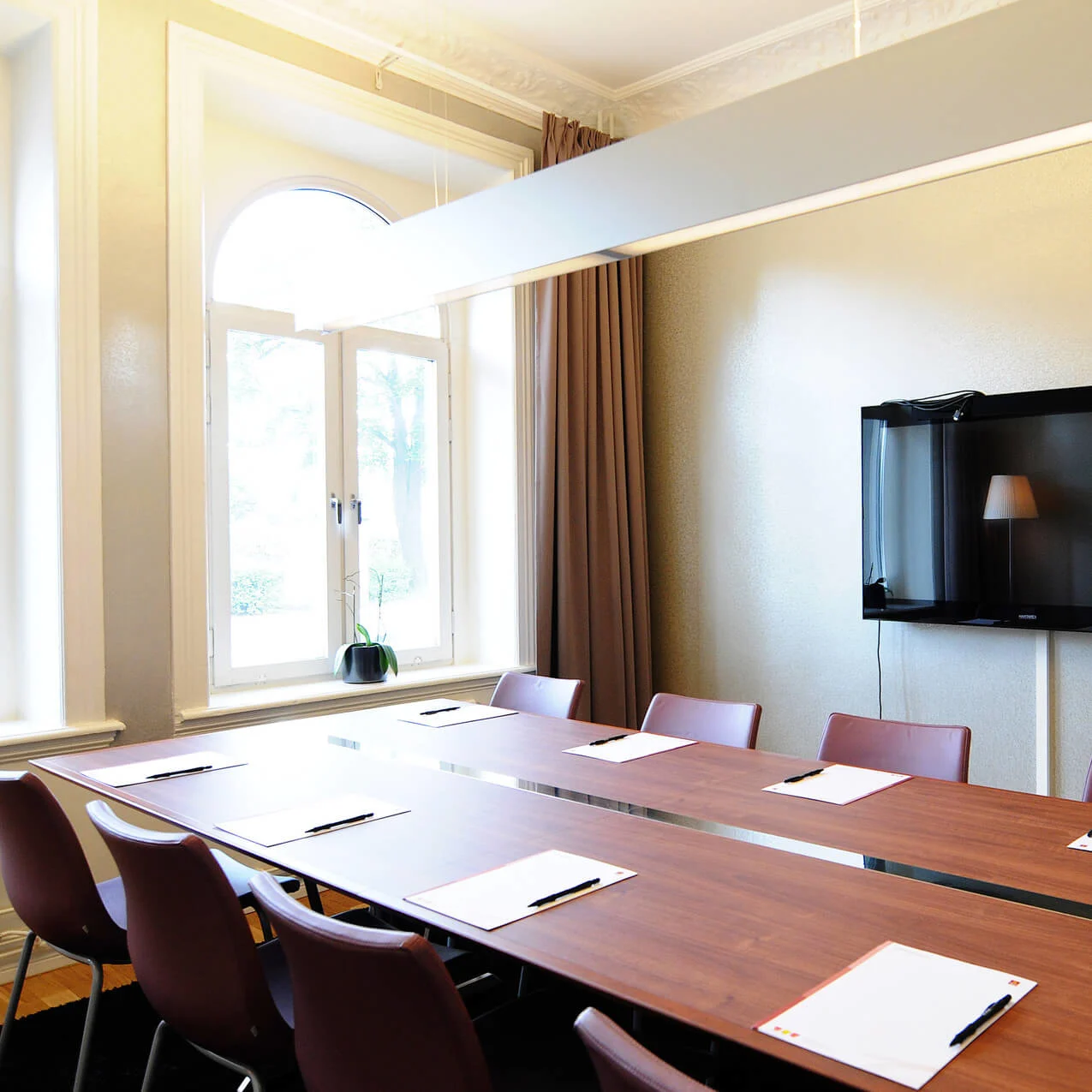 A conference room with a long table, chairs, notepads, and a wall-mounted television. Large window with natural light.