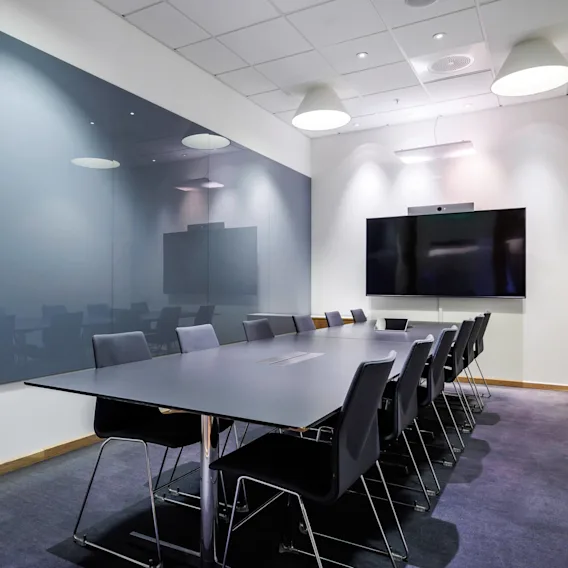 Et konferansebord med svarte stoler er arrangert i et moderne, godt opplyst møterom med en stor veggmontert TV, en glassvegg og hengende lampe.