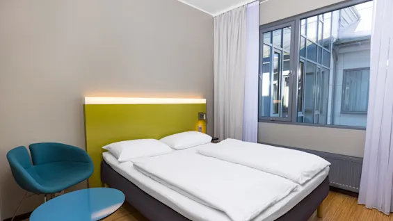 A neatly made bed with white linens stands against a lime green headboard under soft lighting; a blue chair and small round table are beside it, near large windows with sheer curtains.