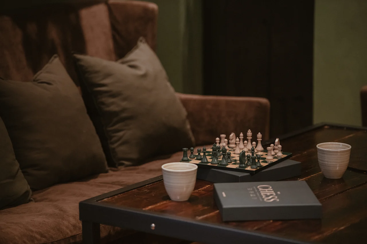Viihtyisä näkymä, jossa shakkilauta on asetettu puupöydälle, kahden kupin ja CHESS-merkityn laatikon kera, mukavan ruskean sohvan vieressä.