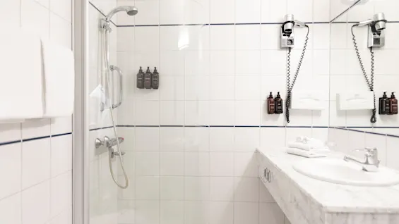 Valoisa ja puhdas hotellihuoneen kylpyhuone, jossa on lasiseinäinen suihku, marmoritasoinen pesuallas ja hygieniatuotteita.