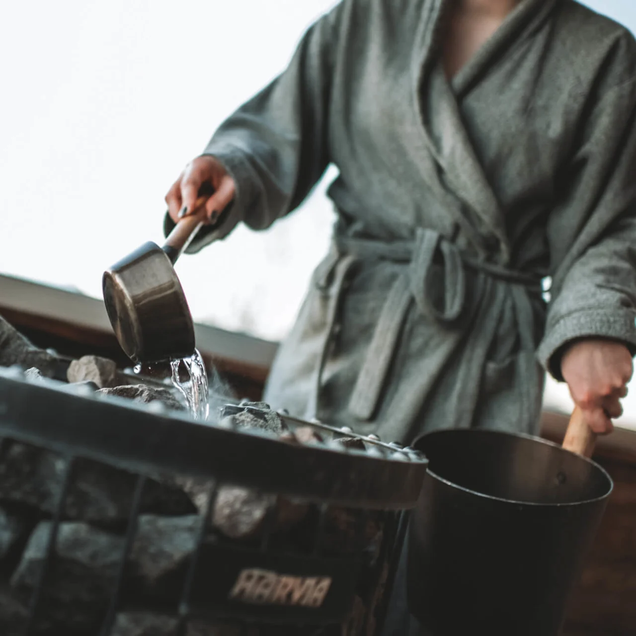 En person i morgonrock häller vatten över heta bastustenar med en metallskopa. Stenarna finns i en metallstruktur med märkningen "HARVIA" inomhus.