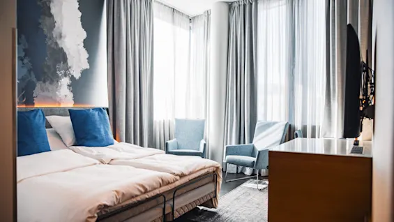 A modern hotel room with a neatly made double bed featuring blue pillows, situated beside abstract art on the wall. Two armchairs face large windows with sheer curtains.