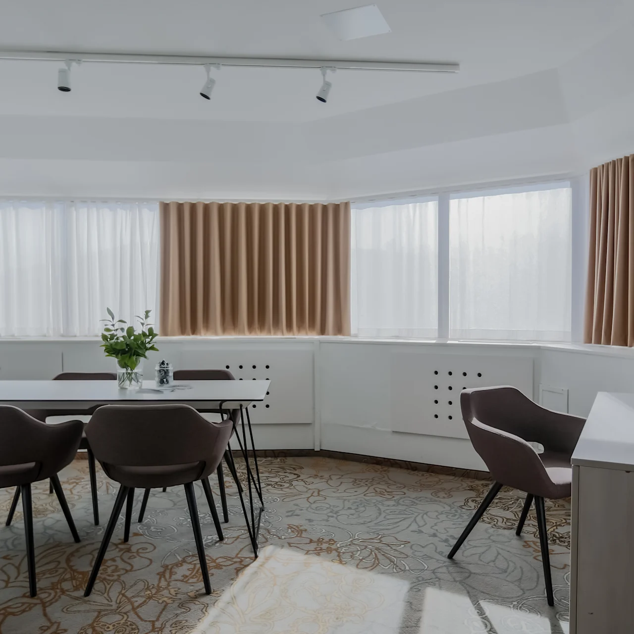 A modern conference room features a table with six chairs, a vase with flowers, and bottles of water. Soft lighting streams through large windows with beige curtains.